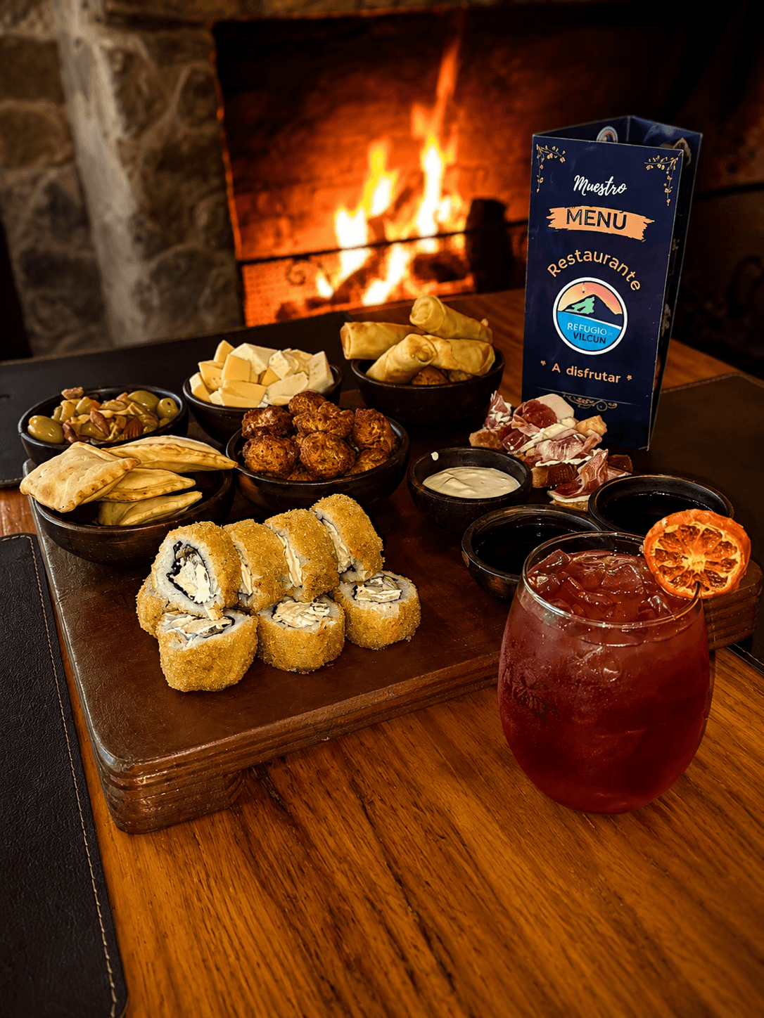 Tabla de picoteo con sushi, quesos, empanaditas y un cóctel de la casa, frente a la chimenea encendida